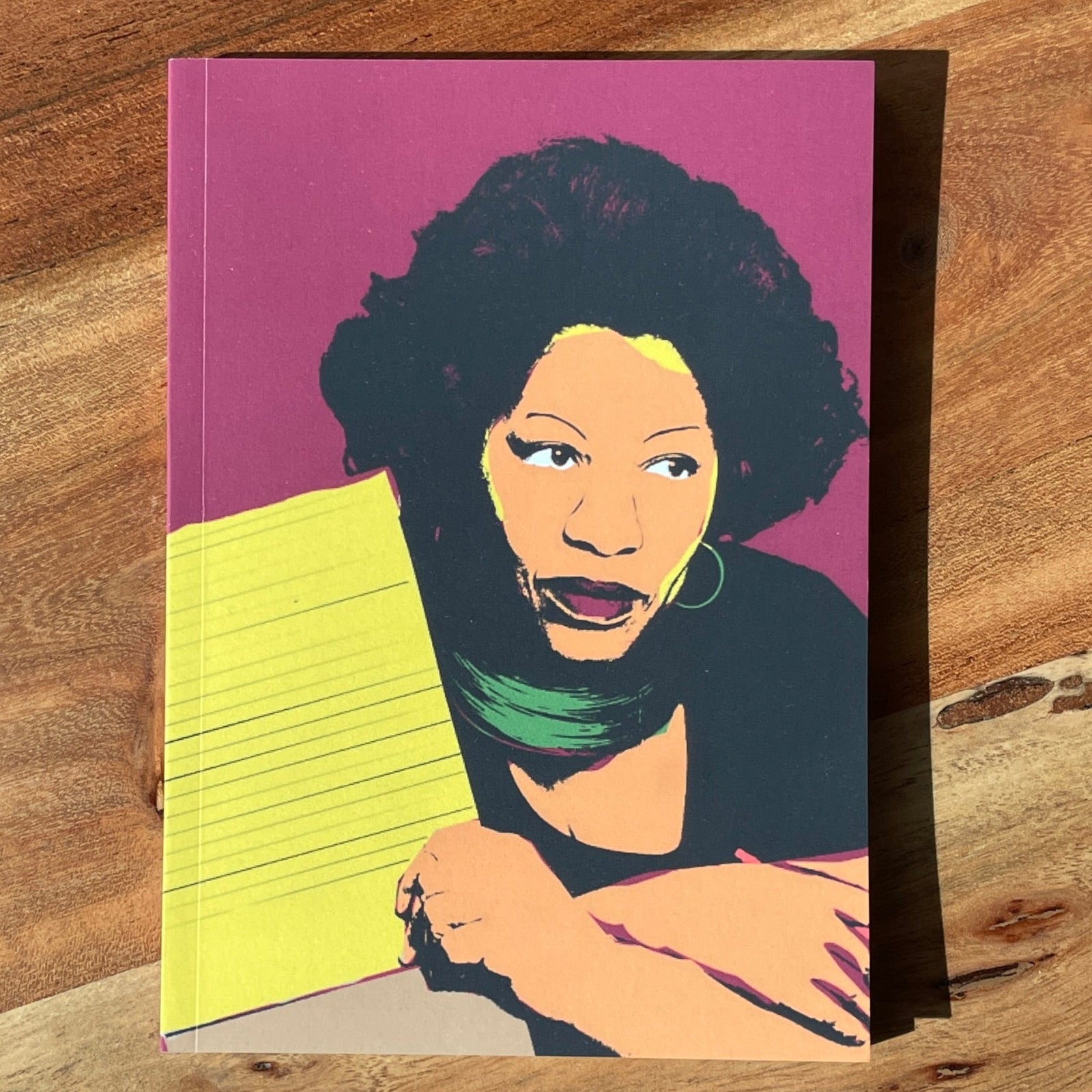 A rectangular art print featuring a stylized pop-art portrait of a Toni Morrison with dark curly hair, a green scarf, and a bright yellow notebook, set against a magenta background. The print is placed on a rustic wooden surface.