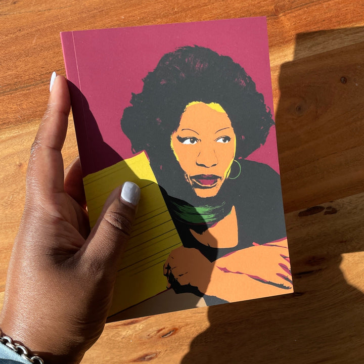 A hand holds a small notebook with a colorful illustrated inspired portrait of Toni Morrison against a warm wooden tabletop in bright sunlight.