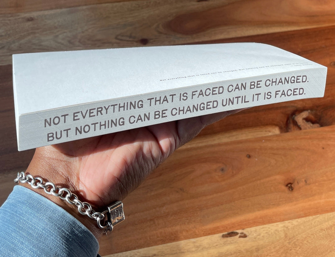 A white rectangular book or notebook with black printed text on its side, held by a hand over a round wooden table. The text reads, 'NOT EVERYTHING THAT IS FACED CAN BE CHANGED, BUT NOTHING CAN BE CHANGED UNTIL IT IS FACED.